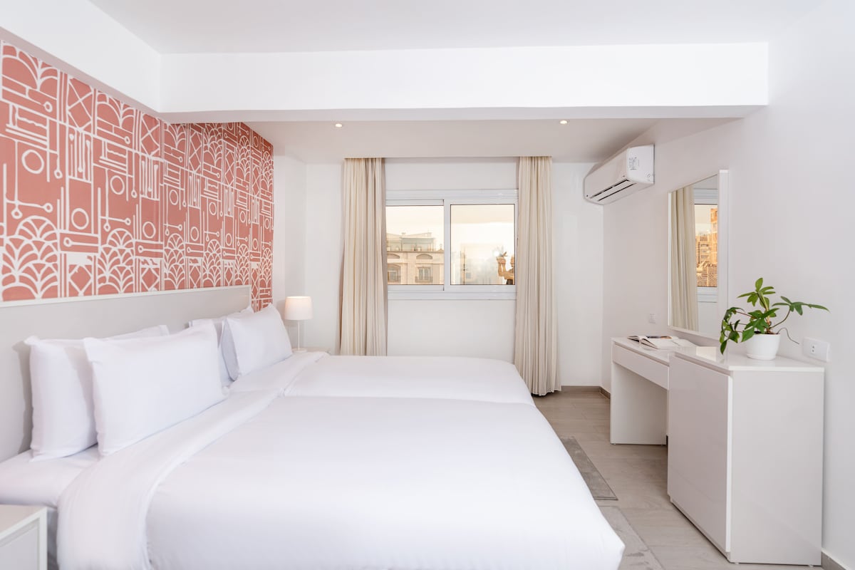 A spacious bedroom is highlighted by a vibrant patterned accent wall. Two large beds are dressed in crisp white linens. Natural light enters through a window, complementing a neat desk and chair setup, alongside a minimalistic dresser. Air conditioning is provided for comfort.