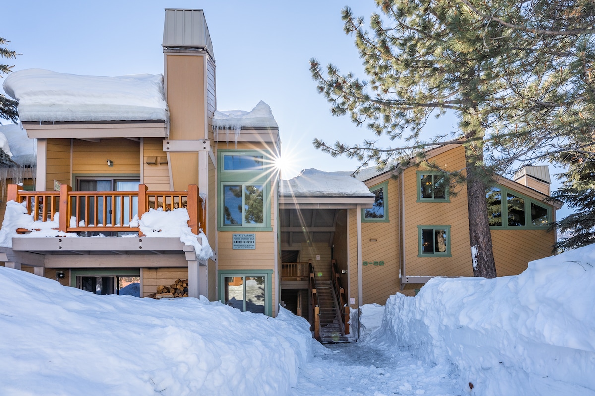 The exterior of a fully remodeled condo is visible, surrounded by snow. Large windows and balconies provide natural light, while a welcoming entrance is framed by trees. Snow-covered paths lead to the entrance, indicating easy access during winter months.