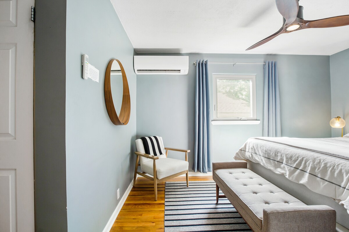 A well-lit bedroom features a spacious bed covered with a white comforter, accompanied by a stylish bench at the foot. A comfortable chair is positioned beside a circular mirror. Light blue walls and a striped rug enhance the room's welcoming ambiance.
