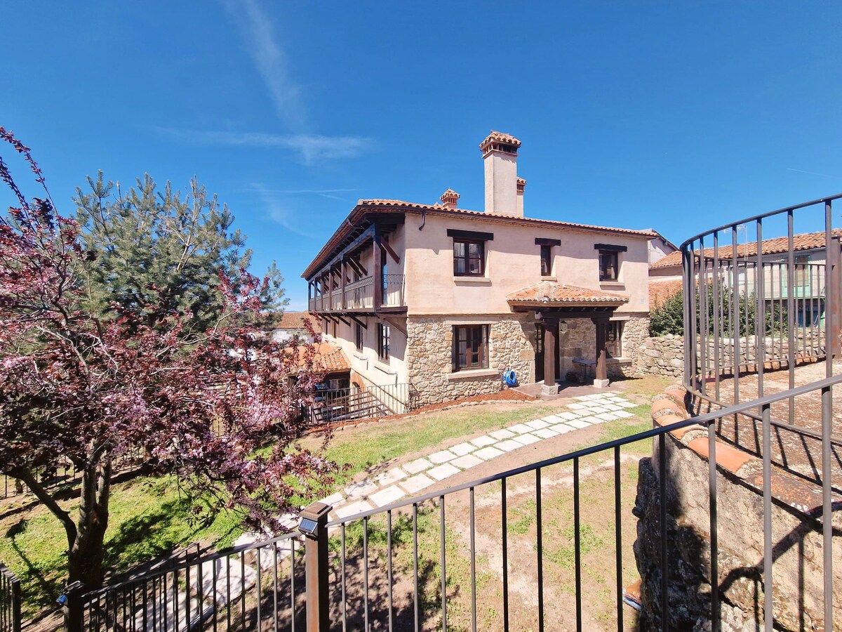 The exterior of a two-story building is displayed, featuring a combination of stone and stucco walls. A pathway made of stones leads to the entrance, framed by a landscaped garden with trees and shrubs. A clear blue sky fills the background.