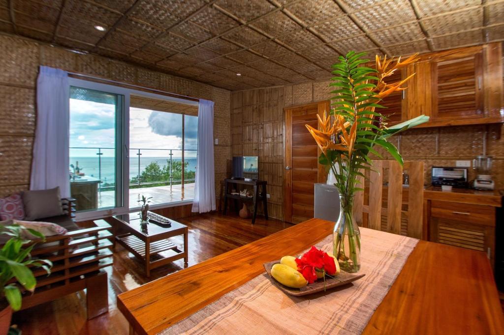 A bright living area showcases polished wooden floors and a bamboo ceiling, creating a unique ambiance. Natural light fills the space through large glass doors leading to the balcony. A dining table is set with fresh fruit and a vase of tropical flowers.