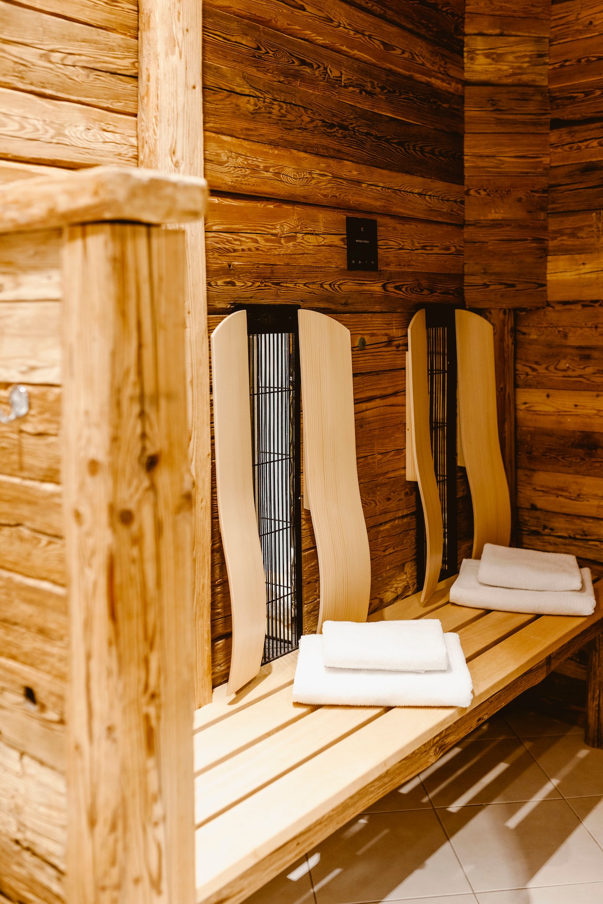 The infrared cabin features natural wood paneling that creates a warm ambiance. Wooden benches are provided alongside folded towels, with a grid design on the wall suggesting a ventilation system. Soft lighting enhances the tranquil environment, inviting relaxation.