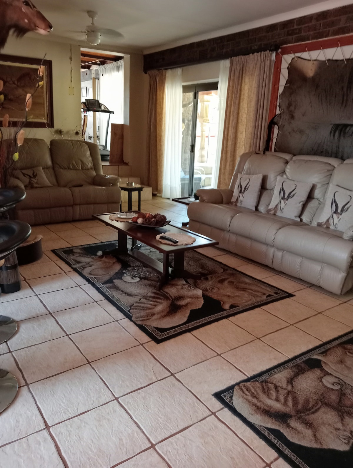 A comfortable living area is presented, featuring two plush sofas and a glass coffee table. Natural light filters through sheer curtains, illuminating the tiled floor and decorative rugs. A doorway leads to an outdoor space, while exercise equipment is discreetly positioned in a corner.