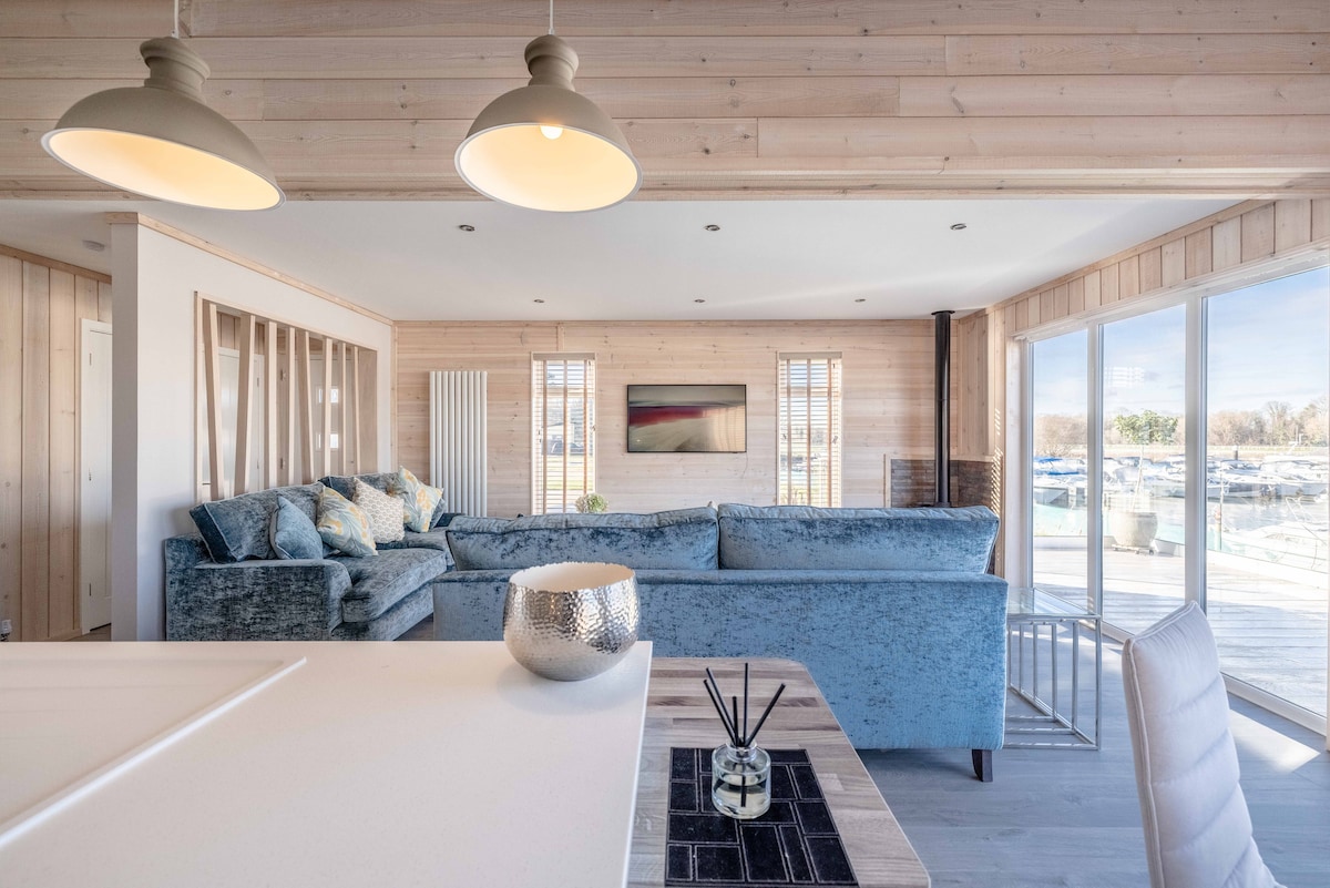 A spacious open-plan living area features a plush blue sectional sofa arranged around a coffee table. Generous windows allow for natural light, revealing views of the marina. A minimalist dining setup is visible in the foreground, with a decorative bowl and elegant aroma diffuser on the table.