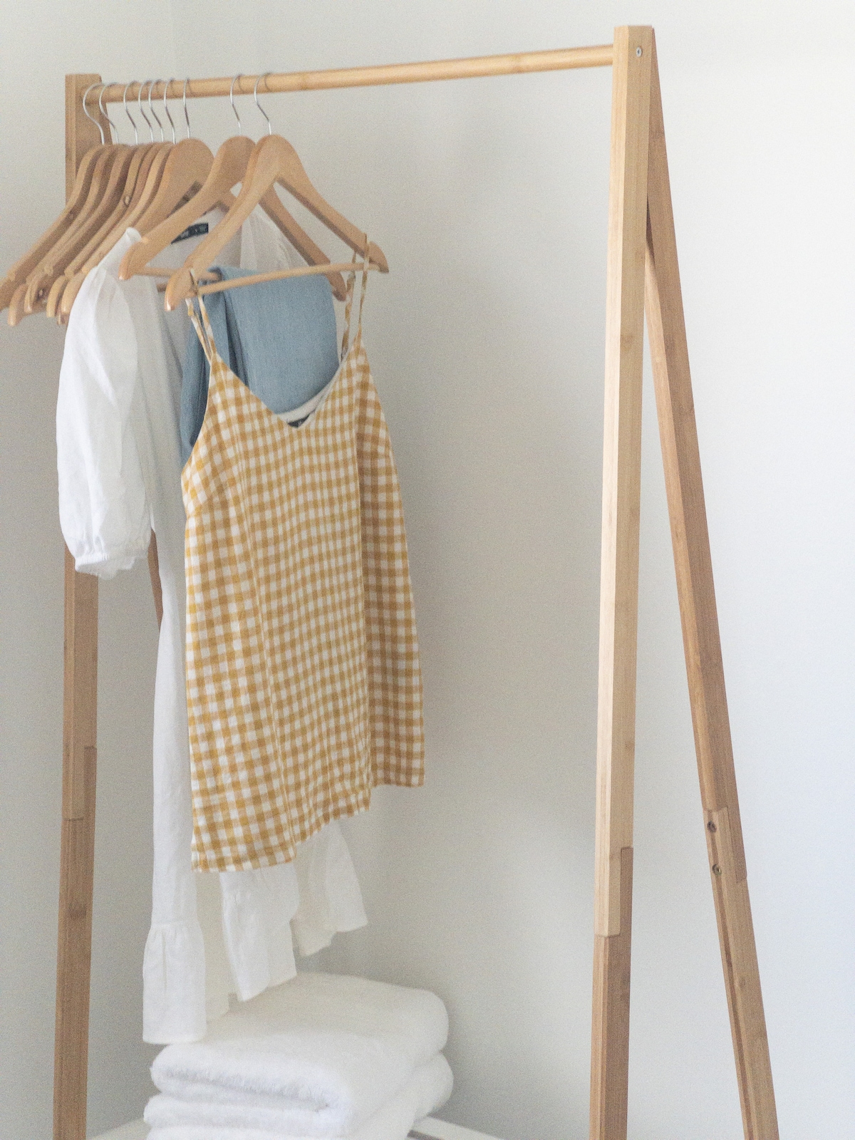 A wooden clothing rack displays several garments, including a light blue top and a white dress with billowed sleeves. A yellow checked sleeveless dress hangs prominently. Neatly stacked white towels are placed below, adding a feeling of organization and accessibility.
