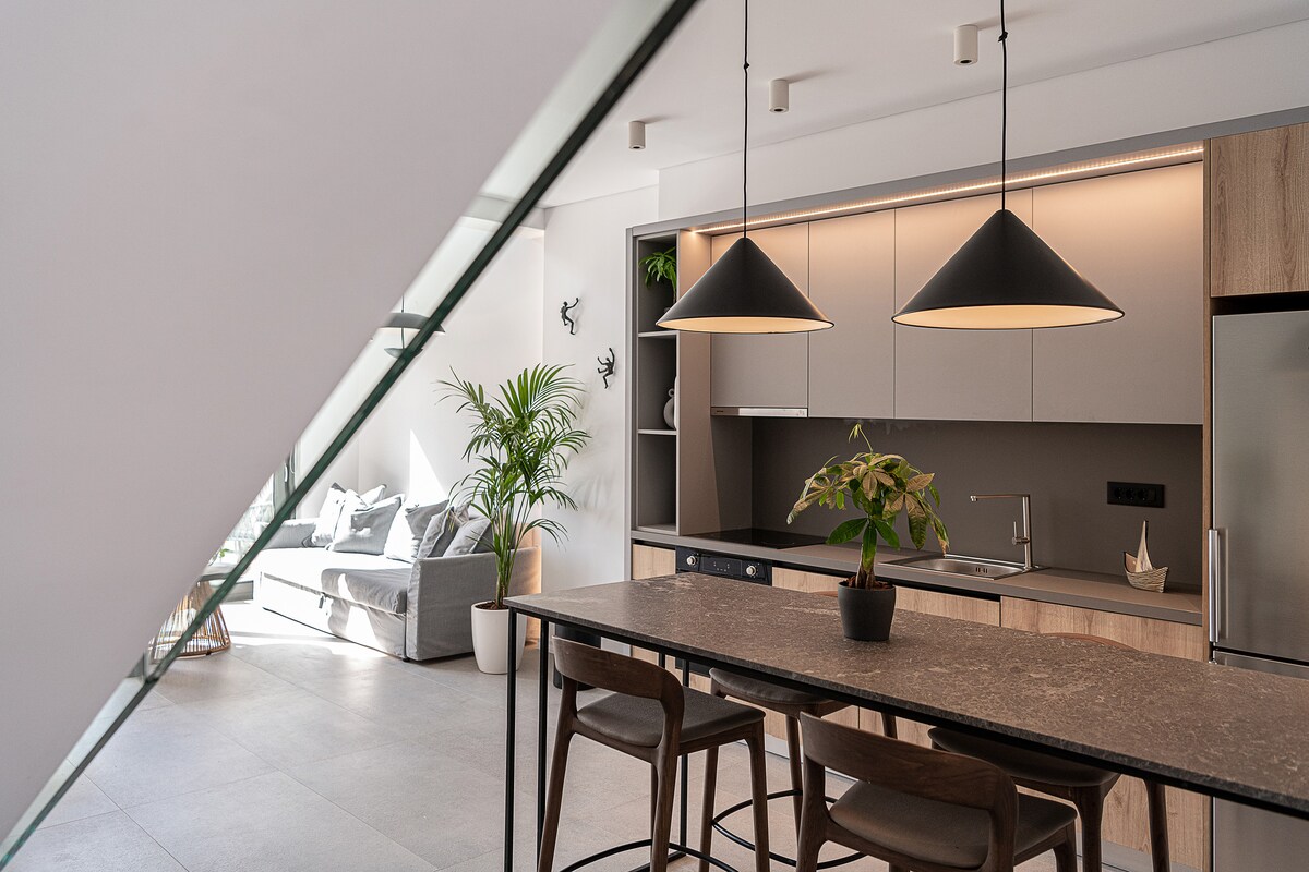A modern kitchen space is highlighted with sleek cabinetry and a spacious island featuring stylish pendant lighting. Plants are positioned near large windows, complementing the minimalistic design. The open-plan layout connects seamlessly to a cozy living area, enhancing the bright and airy atmosphere.