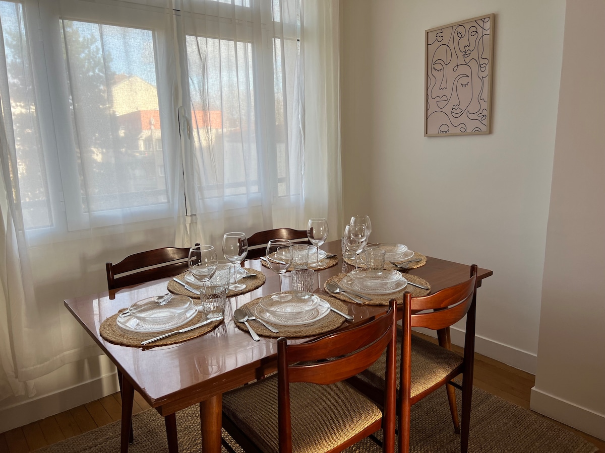 A spacious dining area features a wooden table set for six, complemented by glassware and woven placemats. Large windows allow natural light to fill the room, and a piece of wall art adds a modern touch to the decor.