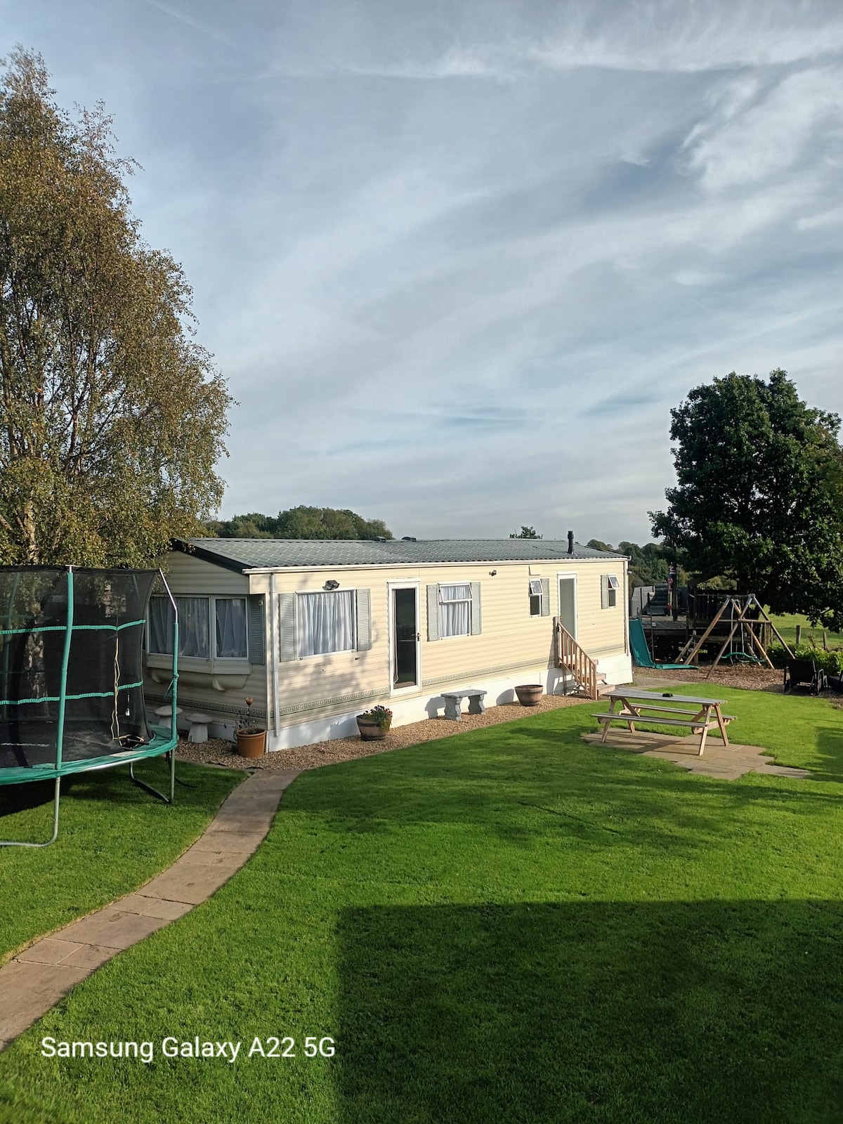 A static caravan is situated in a green yard, surrounded by trees. The exterior features a light yellow façade with windows reflecting the sky. Adjacent to the caravan, a trampoline and a swing set are visible, suggesting a family-friendly environment.