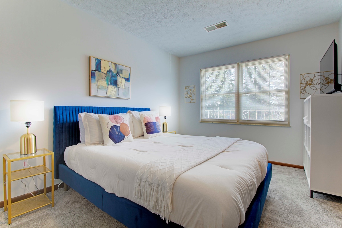 A well-lit bedroom features a spacious bed with plush bedding and decorative pillows. A nightstand with a lamp adds functionality, while a television is positioned on a console against the wall. Two windows allow natural light to brighten the room.