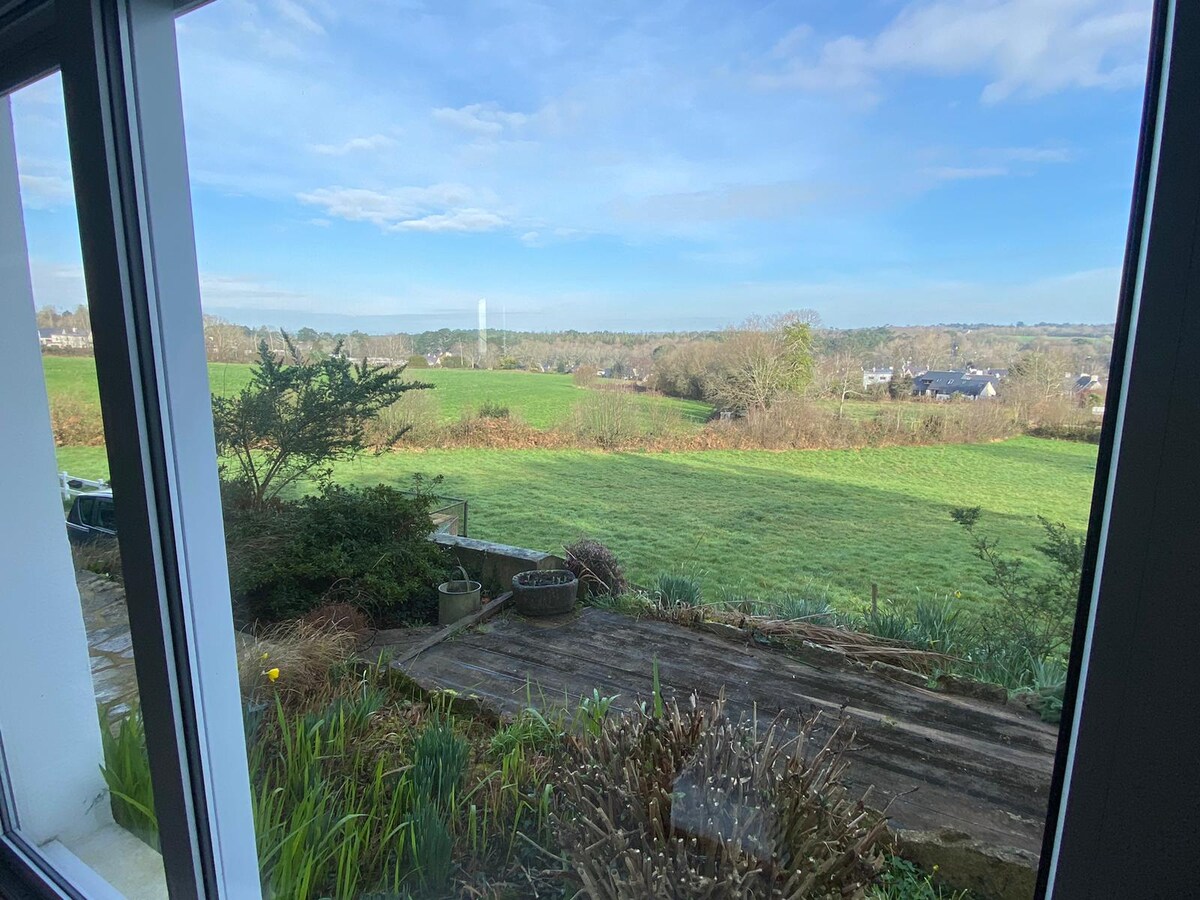 A scenic view of rolling green fields stretches across the landscape. Sunlight brightens the area under a clear blue sky, with a few scattered clouds. The foreground includes garden elements, with shrubs and a stone path leading to the open space.