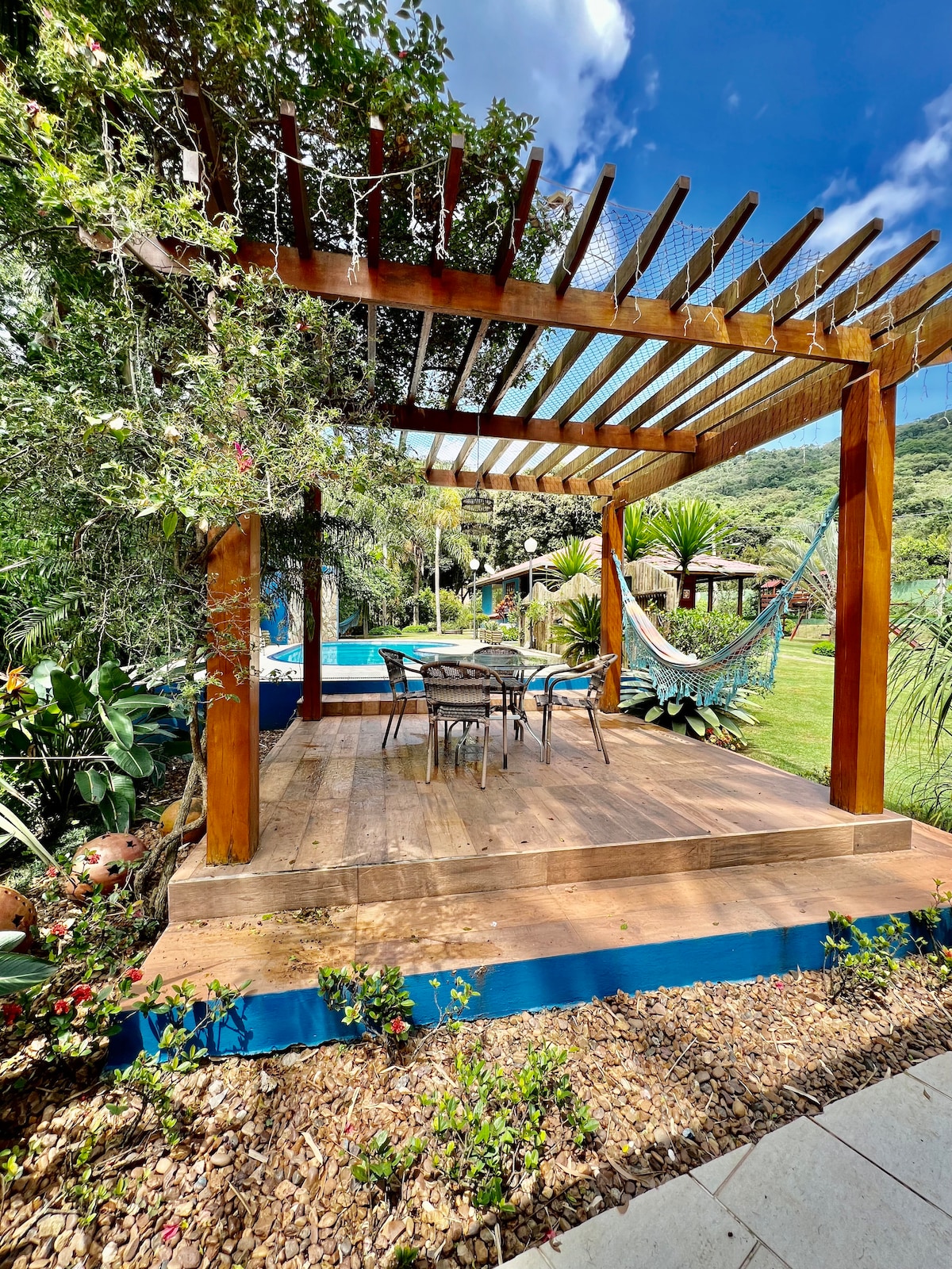 An outdoor shaded area features a wooden pergola supporting a hammock and a dining set with four chairs. The space is surrounded by lush greenery and colorful plants, providing a serene environment near the swimming pool.