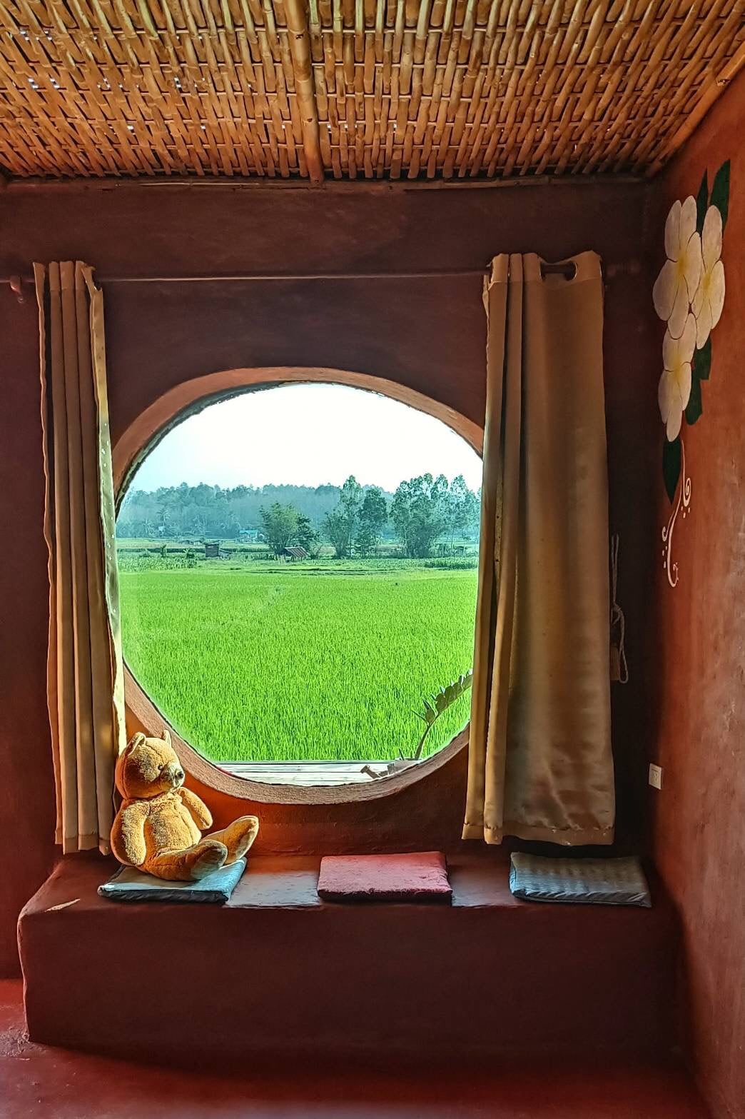 A round window frames a scenic view of lush green rice fields, with soft light filtering through light-colored curtains. A cozy nook features a plush teddy bear seated on a ledge, accompanied by decorative cushions.