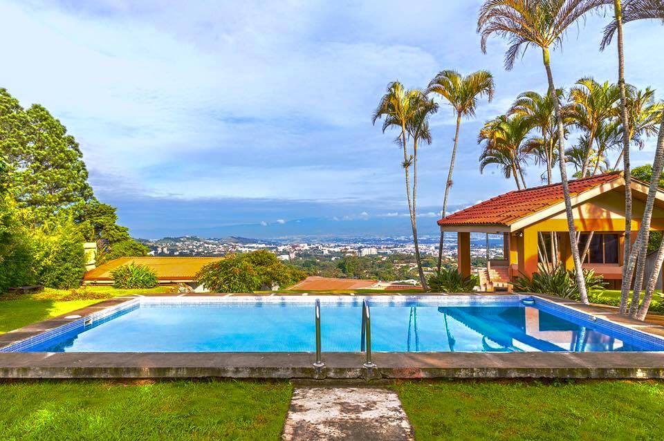 A spacious swimming pool is set against a backdrop of lush greenery and tall palm trees. The inviting water reflects the sky above. A charming house with a terracotta roof stands nearby, providing a view of the surrounding landscape and city below.