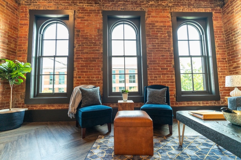 A cozy seating area is showcased with two upholstered chairs and a central ottoman. Large arched windows allow natural light to fill the space, highlighting the exposed brick walls and the warm tones of the flooring. A decorative plant and a small table add to the inviting atmosphere.