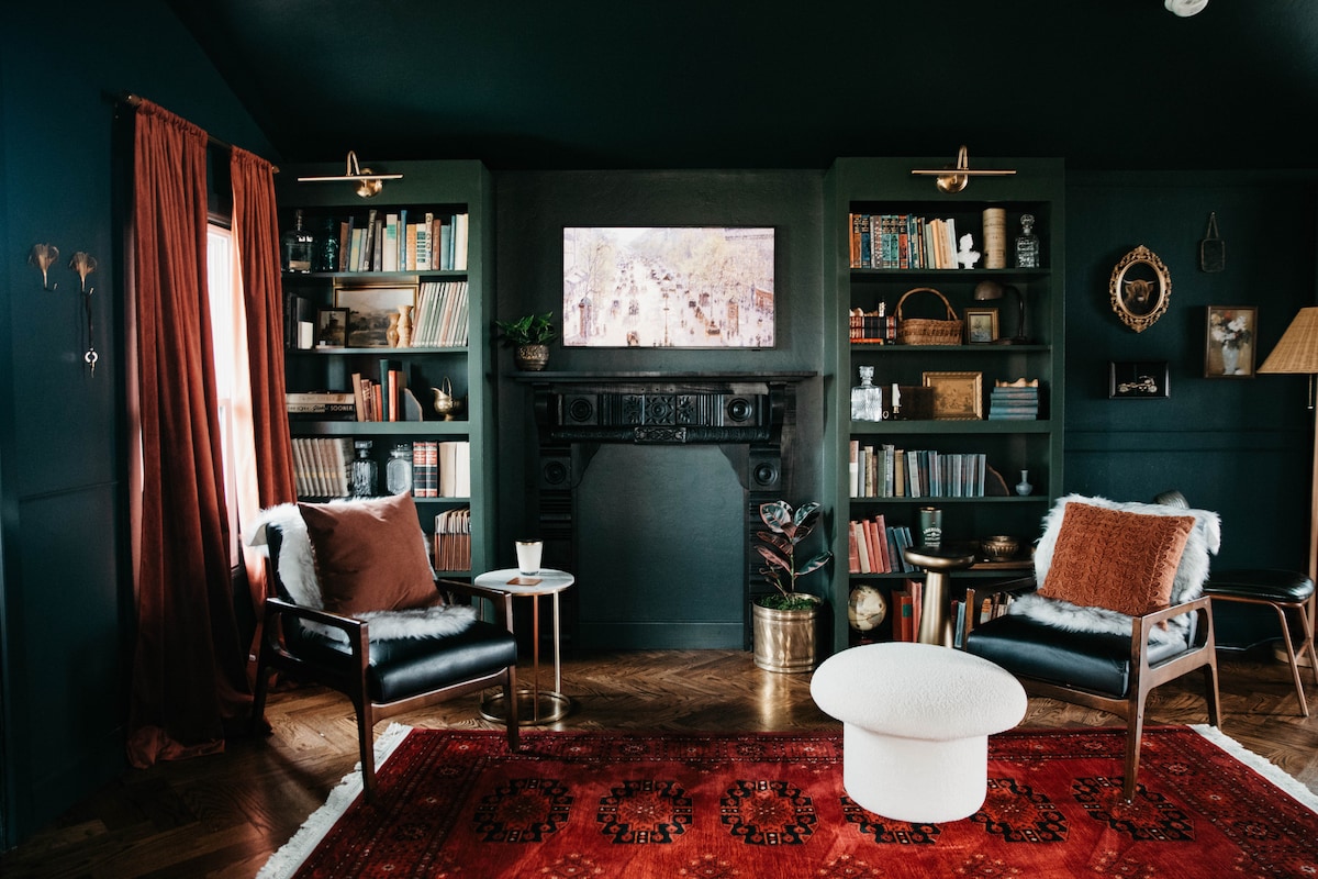 A cozy living area is presented with two black armchairs positioned beside a small table. Dark green bookshelves are filled with books and decorative items. A vintage fireplace serves as a focal point, while a vibrant red area rug adds warmth to the space.
