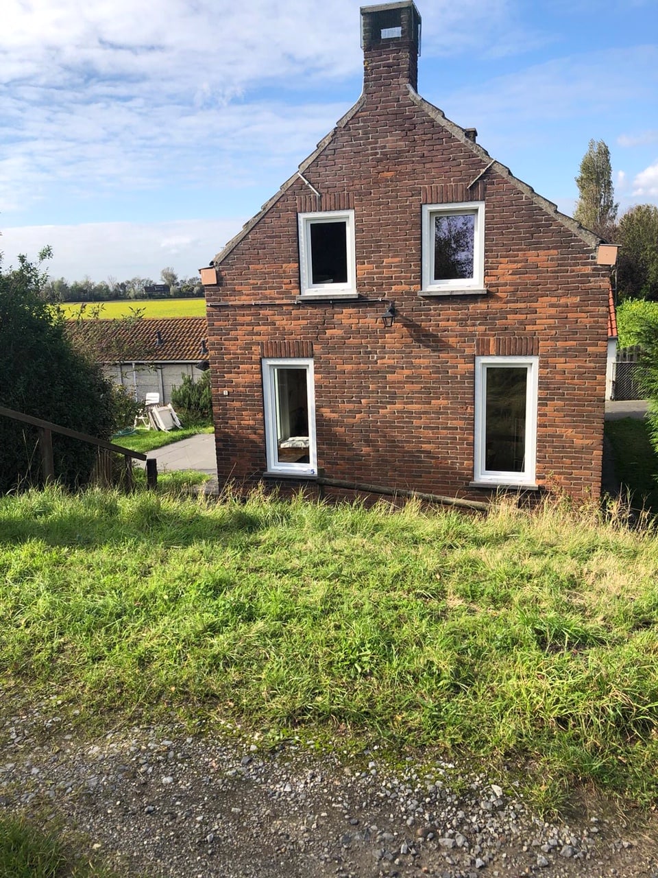 A rustic, two-story brick house features multiple windows, allowing natural light to flood the interior. The landscape showcases a grassy area leading up to the house, with a hint of the surrounding nature visible in the background.