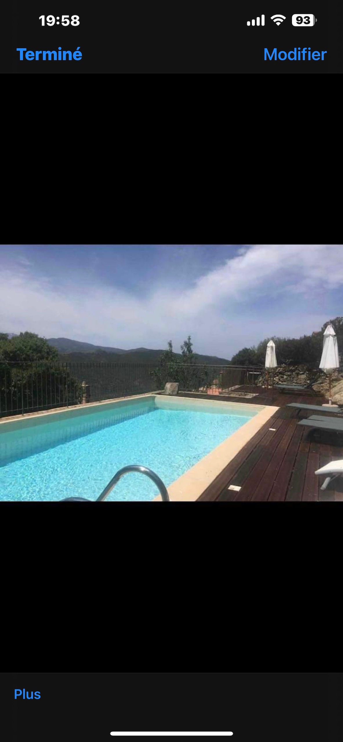 A clear swimming pool is set against a backdrop of rolling hills and a clear sky. Sun loungers are placed on the wooden deck, with umbrellas providing shade. Lush greenery surrounds the area, creating a tranquil environment for relaxation.