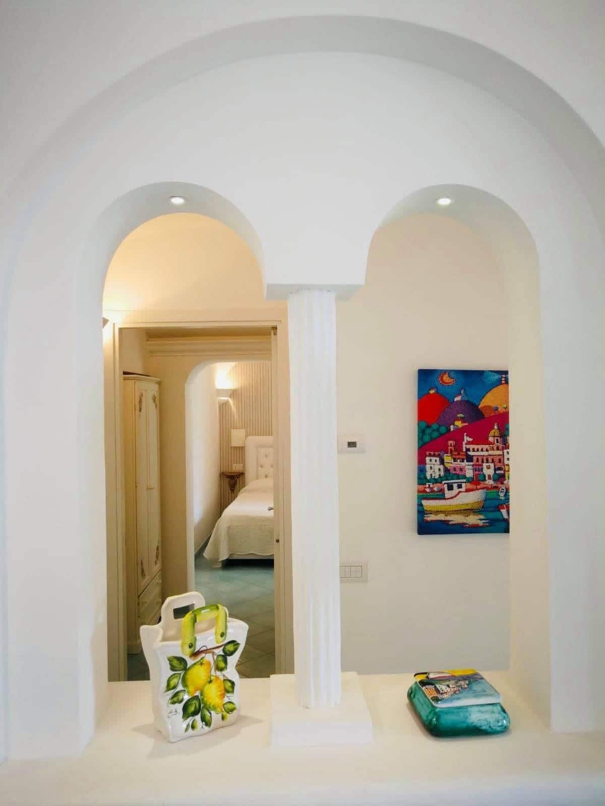 A serene hallway is framed by archways, revealing glimpses of a light-filled bedroom through doorways. A decorative vase with lemons adds a vibrant touch, while a colorful painting on the wall enhances the inviting space. Soft lighting highlights the gentle curves of the architectural features.