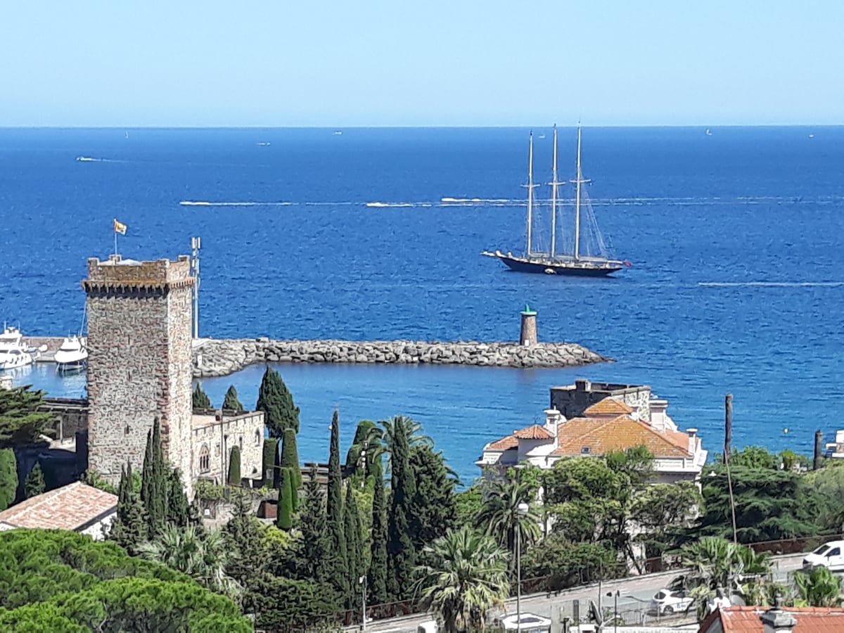 A scenic view showcases the Château de La Napoule against a backdrop of clear blue water. A four-masted sailing ship is seen passing by, with a small lighthouse on the pier and lush greenery surrounding the harbor area.