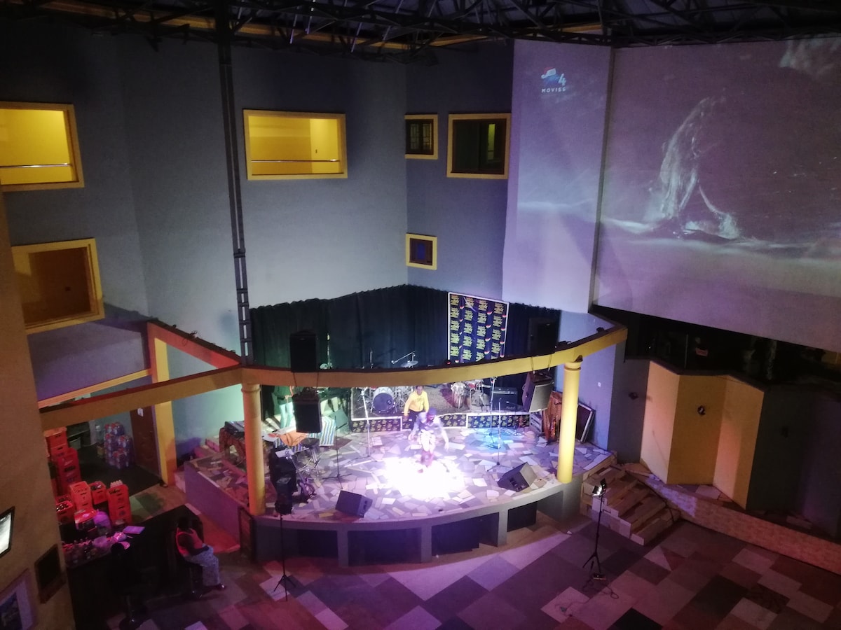 A spacious performance area is highlighted, featuring a circular stage surrounded by seating. Soft lighting enhances the stage where musicians can be seen, with decorative elements and a large screen displaying visual art in the background. Clear paths and open space are present for audience movement.