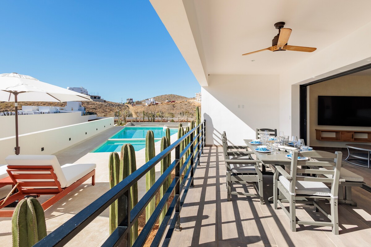 A spacious outdoor terrace is seen, featuring a dining table with seating and a lounge chair. A parasol is positioned for shade, while tall cacti add a natural touch. In the background, a swimming pool glimmers under clear blue skies.