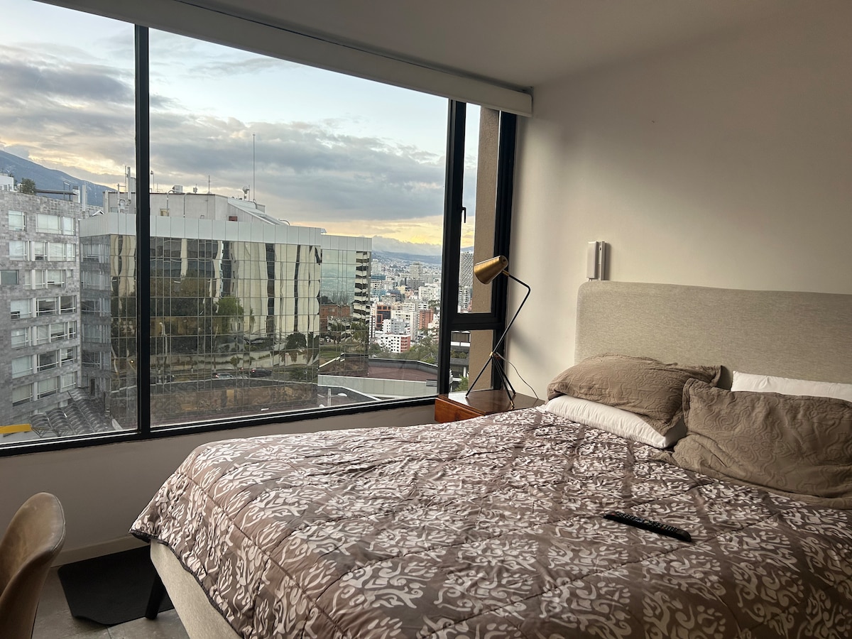 A well-appointed bedroom is situated within a modern high-rise, featuring a spacious bed covered with a patterned blanket. Large floor-to-ceiling windows offer a panoramic view of the skyline, while soft natural light brightens the space.