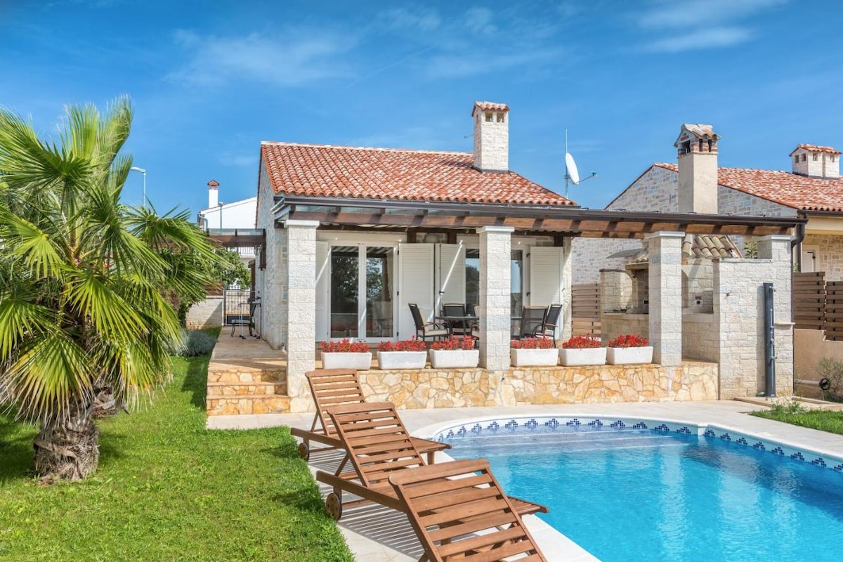 A rustic vacation home features a stone exterior with a tiled roof. A private swimming pool is surrounded by wooden lounge chairs. Spacious terraces are adorned with potted flowers, and large glass doors provide ample natural light. Lush green grass completes the relaxing outdoor environment.