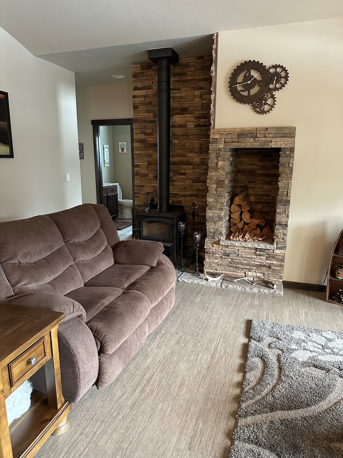 A cozy living area features a plush brown sofa positioned near a stone fireplace, which is stacked with firewood. A wooden side table sits beside the sofa, and a textured area rug covers the floor, enhancing the overall warmth of the space.