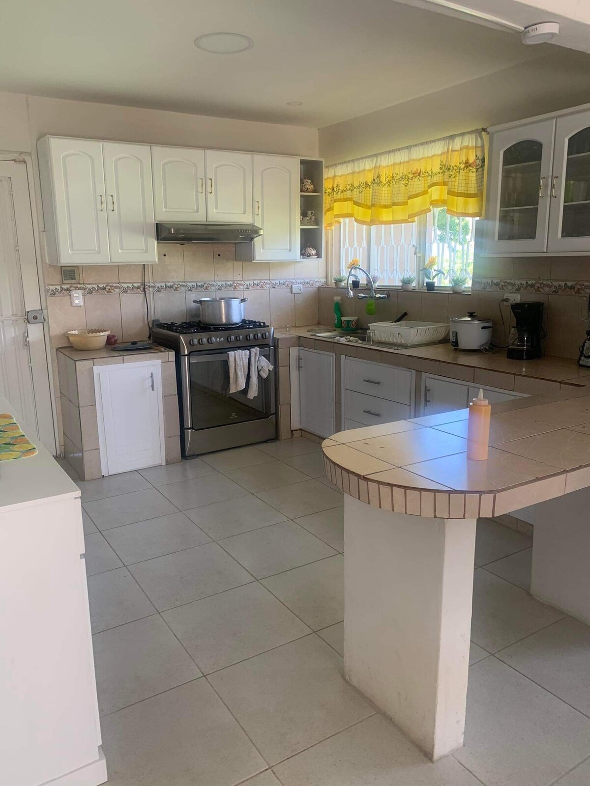 The kitchen features white cabinetry with large windows allowing natural light to fill the space. A gas stove is situated next to a sink, with essential appliances visible. Counter space is available for food preparation, and a small table is positioned nearby.