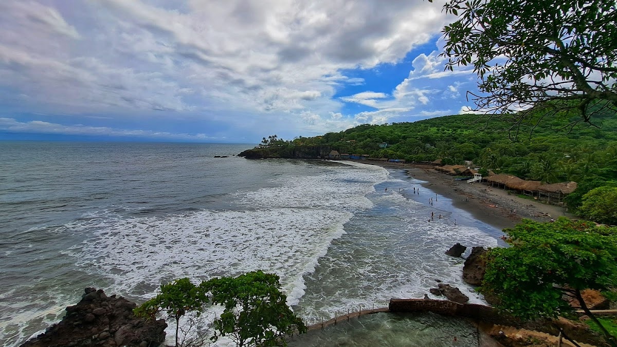 A panoramic view of a serene beach showcases gentle waves lapping at the shoreline. The coastline is bordered by lush greenery, with sandy areas and rocky outcrops visible. A few individuals can be seen enjoying the water, and a cluster of thatched-roof structures is positioned along the beach.