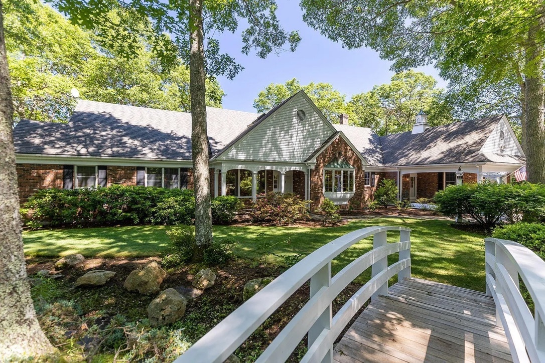 A charming exterior view of a single-story brick home is surrounded by lush greenery and mature trees. A wooden footbridge leads to the entrance, providing a welcoming pathway. Sunlight filters through the leaves, highlighting the inviting facade characterized by large windows.
