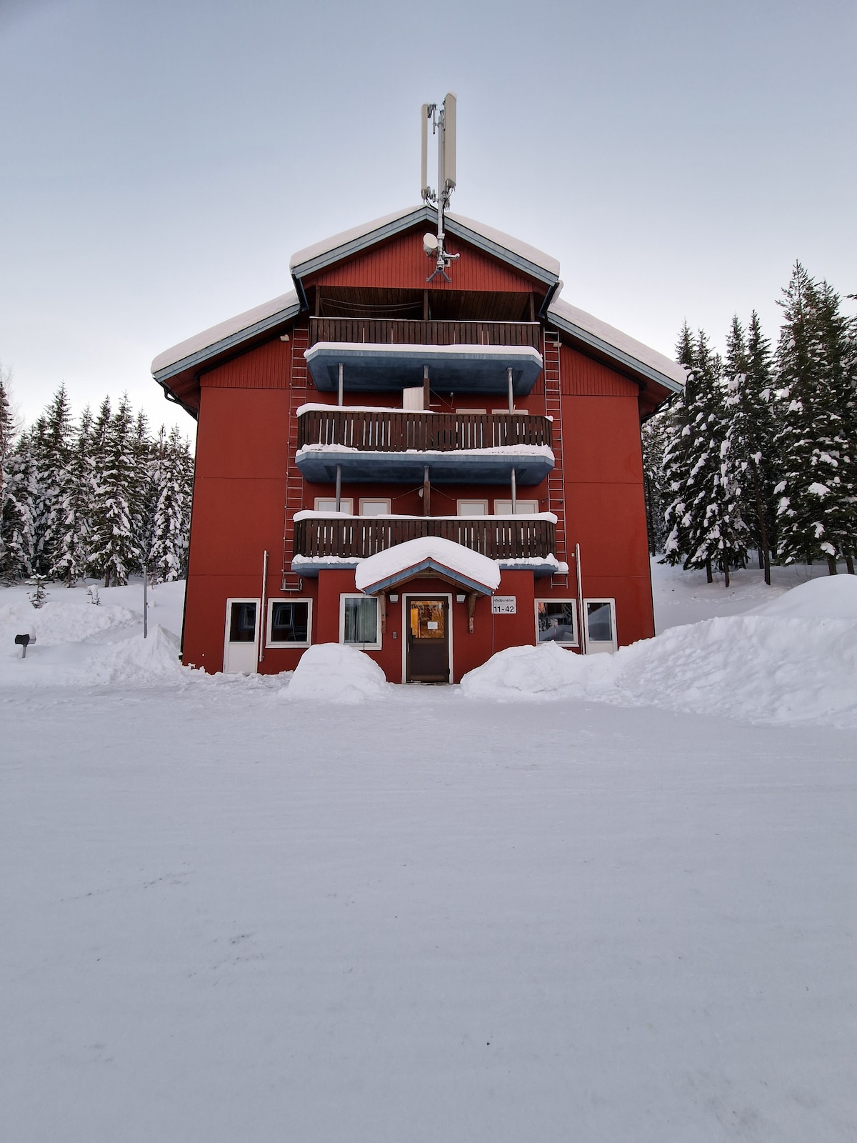 A three-story red building is surrounded by snow-covered grounds, with several balconies visible on the upper levels. The entrance has a welcoming door flanked by large windows. Tall evergreen trees can be seen in the background, contributing to the winter landscape.