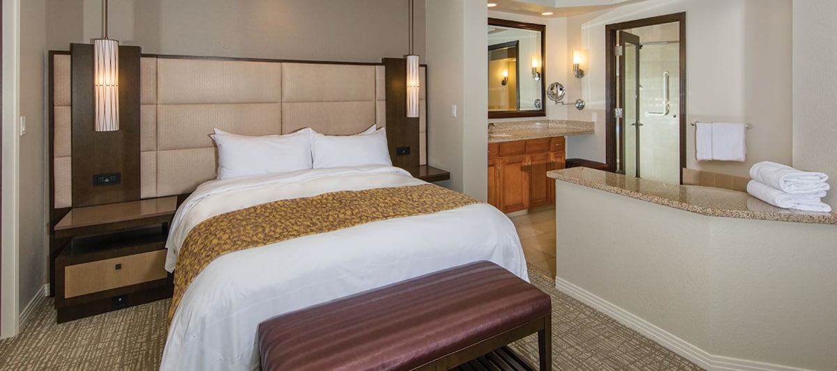 A spacious one-bedroom Villa is presented with a large bed featuring plush linens. Nightstands with integrated lighting and a stylish bench add functionality. An open layout reveals a well-equipped kitchen area in the background, while the bathroom is distinguishable by granite countertops and tasteful fixtures.