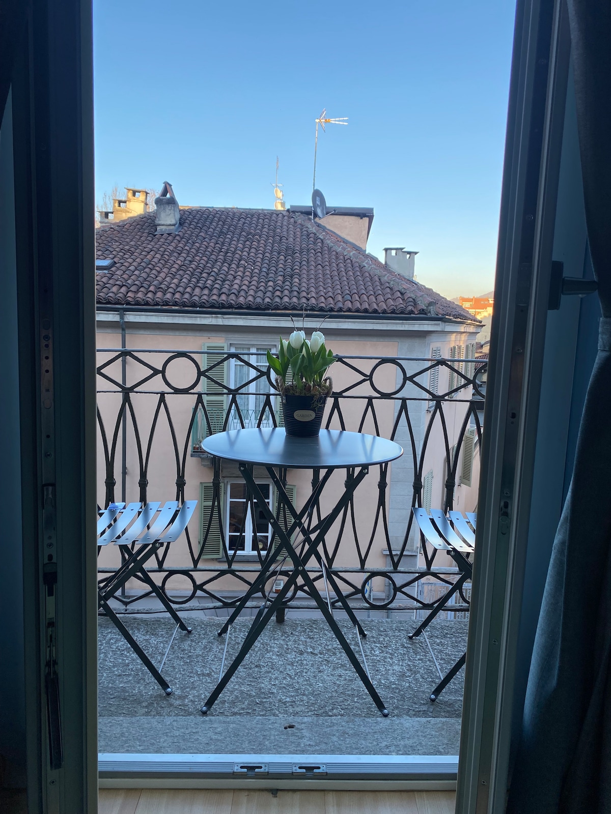 A small balcony is visible through open double doors, featuring a round table and two foldable chairs. A potted plant sits on the table, while the backdrop showcases the rooftops of adjacent buildings under a clear blue sky.