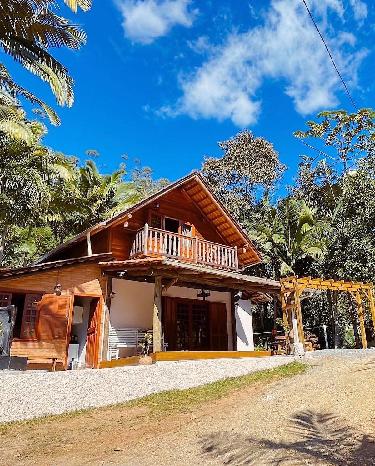 A charming wooden cabin surrounded by lush greenery features a steep, thatched roof and spacious balcony. Sunlight filters through the trees, enhancing the natural setting. A gravel pathway leads to the entrance, inviting visitors to explore the serene landscape.