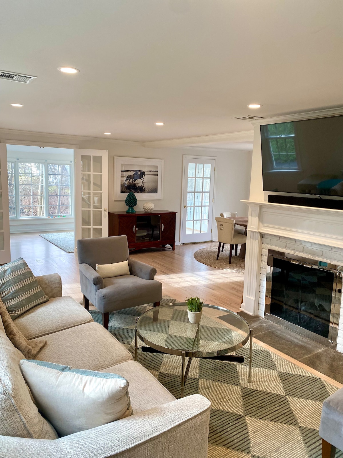 A light-filled living area is presented with a comfortable sofa and an armchair, complemented by a round glass coffee table. A flat-screen television is mounted above a marble fireplace. French doors lead to a serene outdoor space, with a small dining area in view.