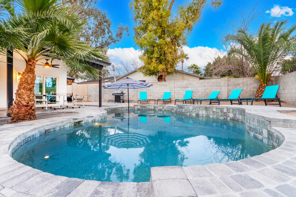 A heated swimming pool is surrounded by lounge chairs in vibrant blue, with palm trees providing shade. The stone patio is designed for relaxation, and a grill area is visible in the background, creating an inviting outdoor space for guests.