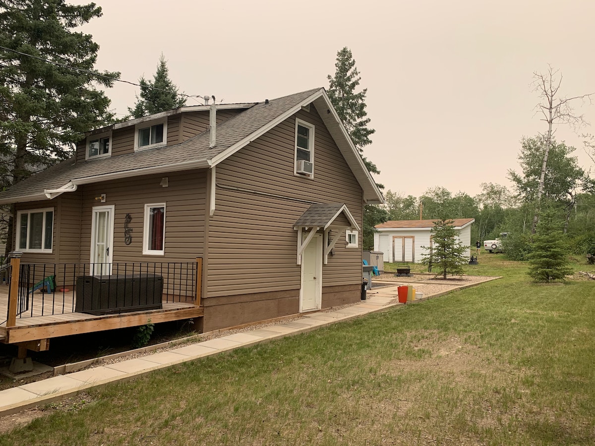 The side view of a newly renovated cabin is presented, featuring a wooden deck with seating and a spacious lawn area. A pathway leads to the entrance, with trees lining the property, and additional structures visible in the background.