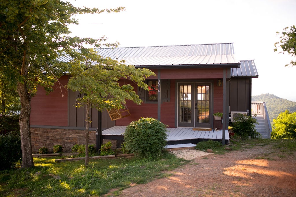The exterior of a modern home is depicted, featuring a sloped metal roof and a mix of wood and stone finishes. A front porch is visible with a swing, while landscaping includes greenery and stone pathways. A view of the mountains is seen in the background.