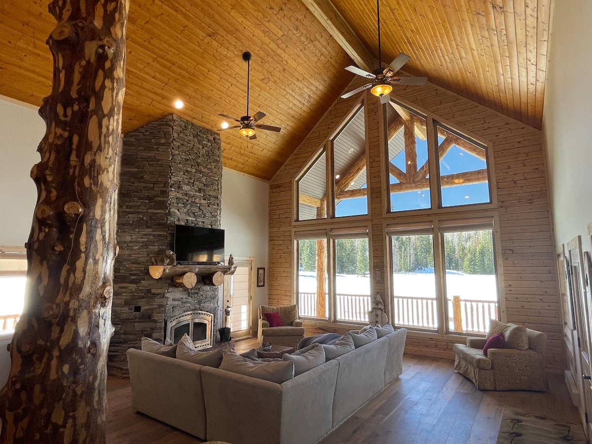 The spacious great room features a high vaulted ceiling with wooden beams, large windows overlooking a snowy landscape, and a stone fireplace with a mounted television above. A comfortable sectional sofa and an accent chair are positioned for relaxation.