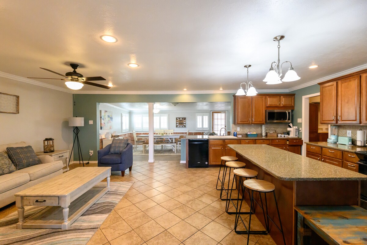 A spacious kitchen occupies the center of the image, featuring a large island with seating for three. Cabinets in warm wood tones complement the light granite countertops. The adjacent living area is visible, showcasing a comfortable sofa and additional seating, with soft lighting enhancing the inviting atmosphere.