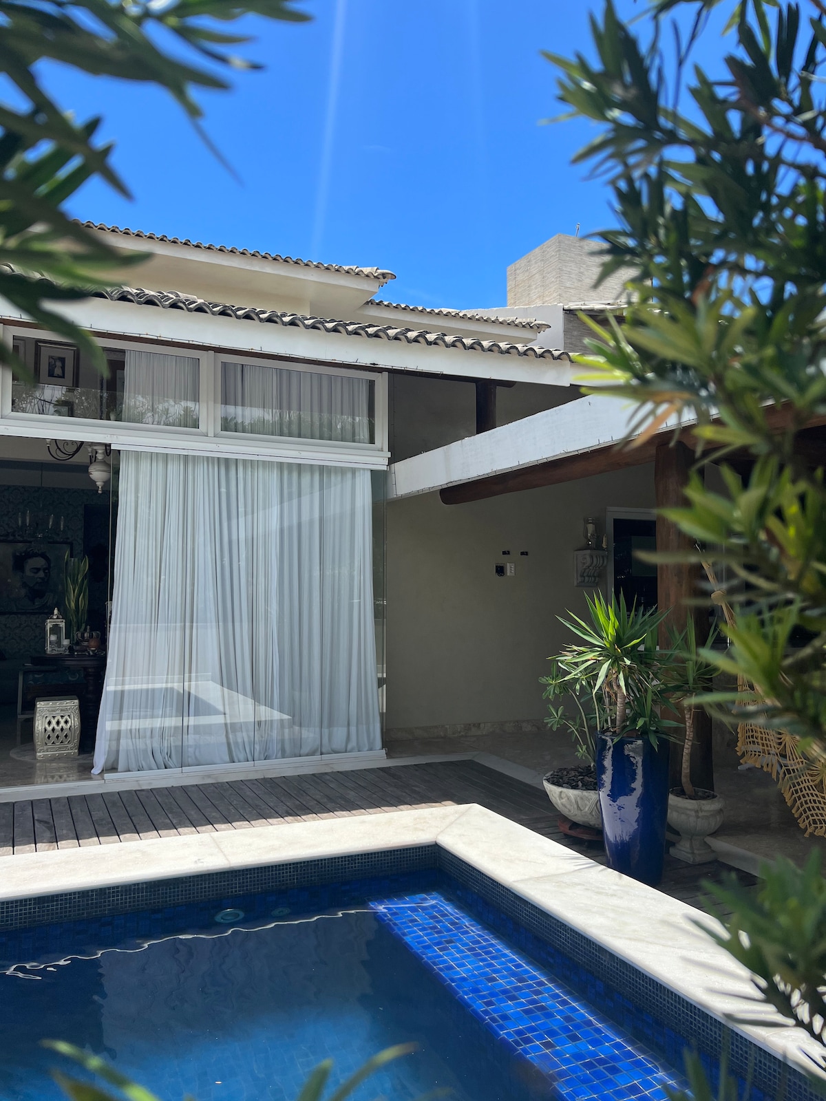 A refreshing swimming pool is framed by a wooden deck and lush greenery. The sunlit patio area features a spacious home with large windows, allowing plenty of natural light. A serene atmosphere is enhanced by the soft flow of sheer curtains in the background.