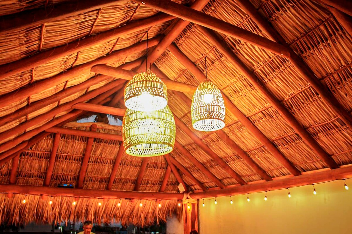 A straw thatched roof is supported by wooden beams, creating a warm, inviting space. Three woven pendant lights provide soft illumination, complementing the ambiance. Below, string lights are mounted along the walls, enhancing the tranquil setting.