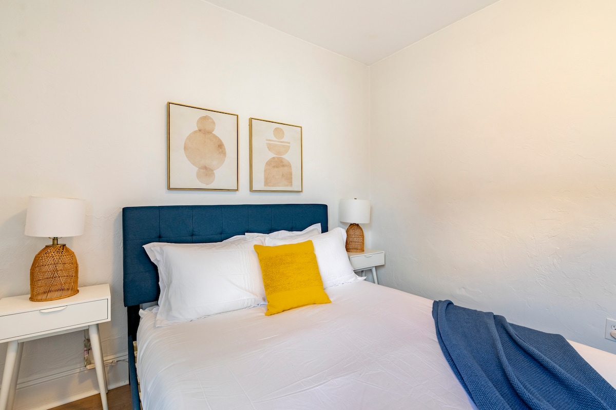 A modern bedroom showcases a spacious bed with white linens and a blue upholstered headboard. Two bedside tables with woven lamps sit on either side. Above the bed, two framed art pieces add a touch of decor. A soft throw blanket rests casually on the bed.