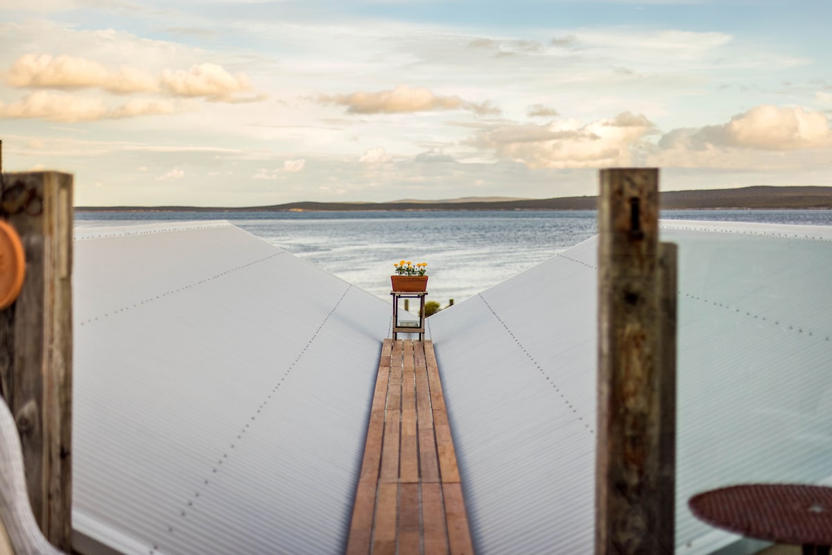 A wooden walkway extends towards a serene body of water, framed by slanted metal roofs. A small table adorned with flowers is positioned at the walkway's end, serving as a focal point against the expansive view of the horizon.