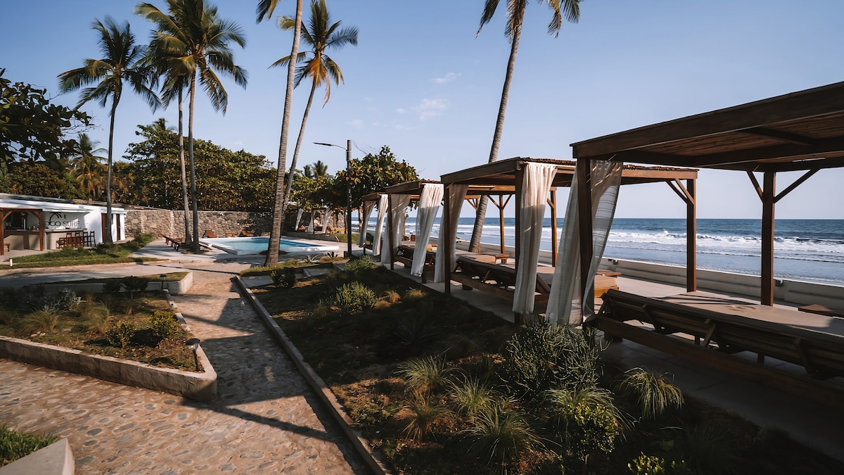 A serene outdoor area is shown, featuring private cabanas arranged along a pathway. Lush greenery surrounds the space, while palm trees sway gently in the breeze. The tranquil ocean is visible in the background, with soft waves lapping at the shore under a clear blue sky.