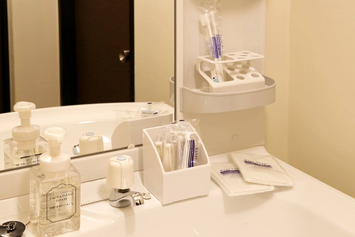A clean and organized bathroom countertop is featured, displaying various toiletries including liquid soap, toothbrushes, and disposable cups. Wrapped items are neatly arranged beside a small tissue box, and white towels are folded nearby, providing essential amenities for a comfortable stay.