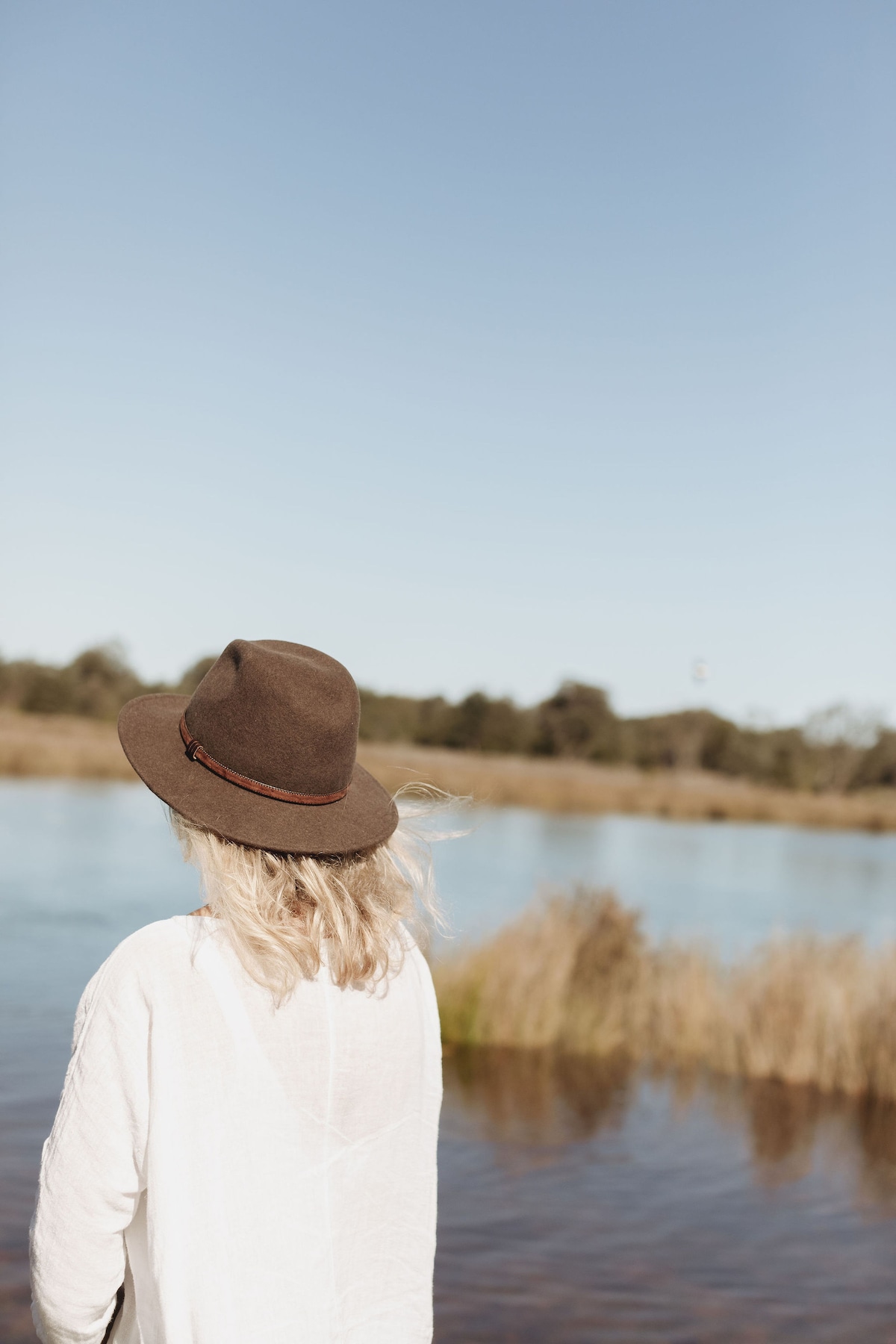 A woman with long, light-colored hair is seen from behind, wearing a brown hat. She stands near a calm body of water, which reflects the clear blue sky. Tall grasses and shrubs frame the serene landscape, creating a peaceful natural setting.