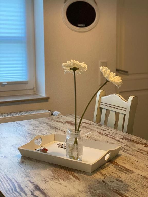 A rustic wooden table is complemented by a simple arrangement of two white flowers in a glass jar placed on a light-colored tray. A single candle is also visible on the tray, with natural light filtering in through a nearby window.