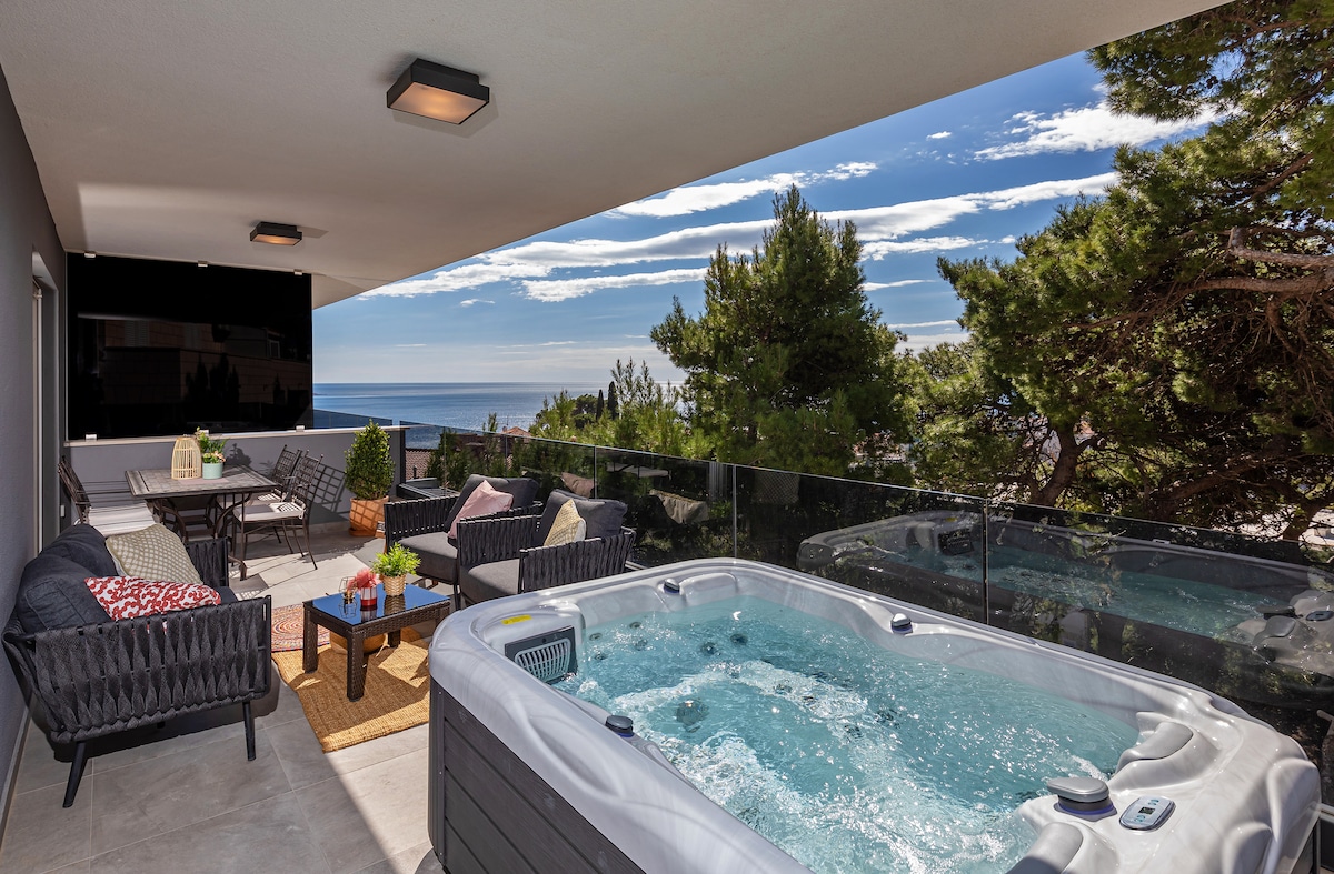 A spacious balcony features a welcoming jacuzzi, surrounded by potted plants and comfortable seating. Sea views extend beyond a glass railing, with trees providing a touch of greenery. The bright sky adds to the serene atmosphere of the outdoor area.